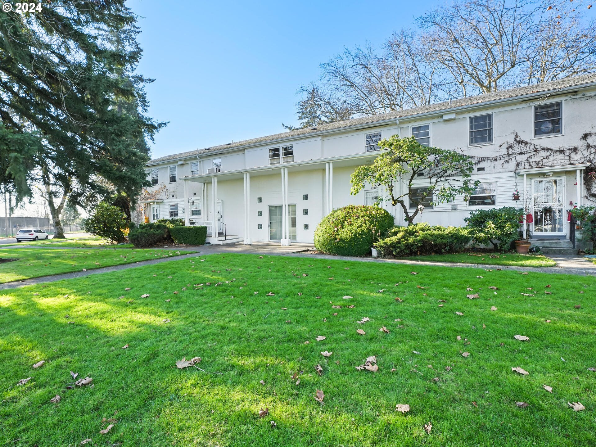 a view of a white house in front of a big yard plants and large trees