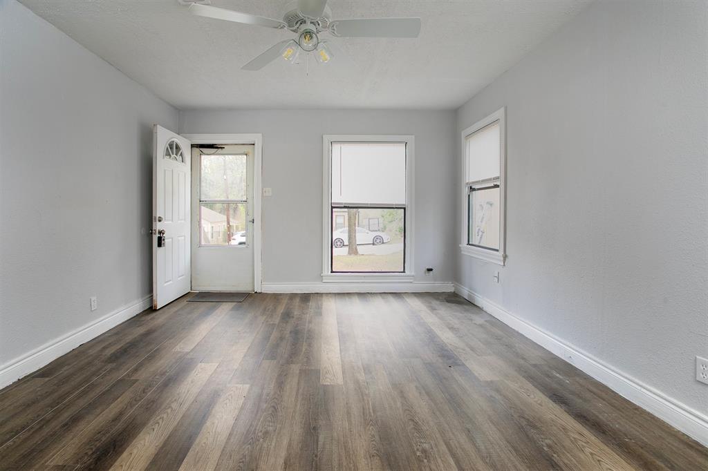 This is a bright, empty room featuring a fresh, neutral color scheme, modern laminate flooring, ample natural light from windows, and a ceiling fan, with a view of the front door leading outside.