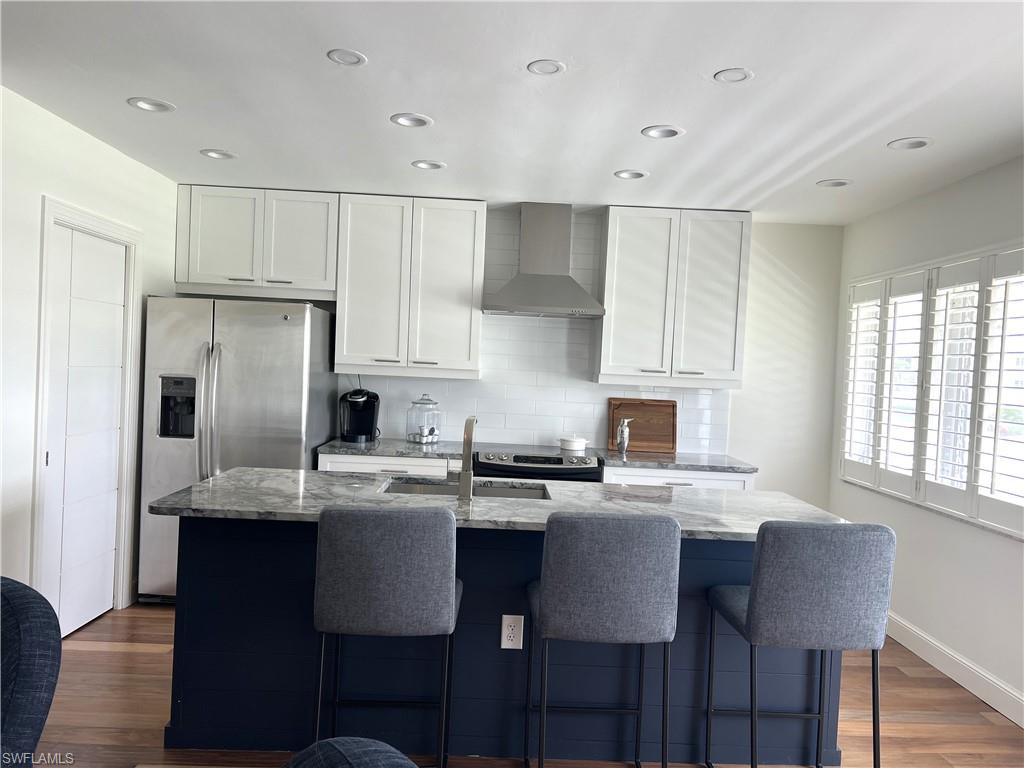 Kitchen featuring a center island with sink, light stone countertops, wall chimney range hood, and white cabinets