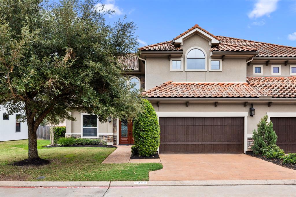 This is a charming two-story house with a Mediterranean-style design. It features a tile roof, arched windows, and a double garage. The front yard is landscaped with a large tree and shrubs, giving it a welcoming curb appeal.