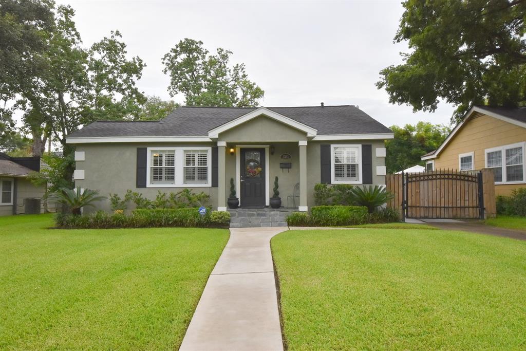 Charming single-story home with a neat gray exterior, black shutters, and an inviting covered entryway. Features a well-maintained lawn, a driveway leading to a secure gate, and mature trees offering shade.