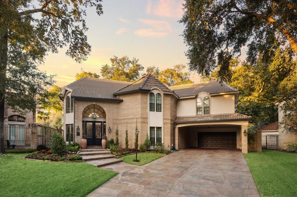 This elegant two-story home features a brick exterior, tile roof, and a spacious two-car garage. Landscaped with well-maintained greenery, it offers a welcoming entrance with a path leading to an arched front door, accented by outdoor lighting and flanked by large trees, giving the property a serene and established setting.