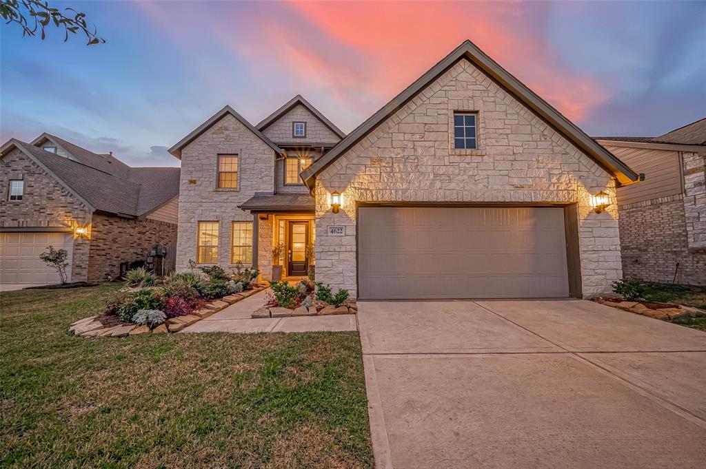 This photo showcases a charming two-story home with a stone exterior and a spacious two-car garage. The well-maintained front yard features a landscaped walkway, and the home is warmly lit, creating an inviting atmosphere under a beautiful sunset sky. Builder Lennar.