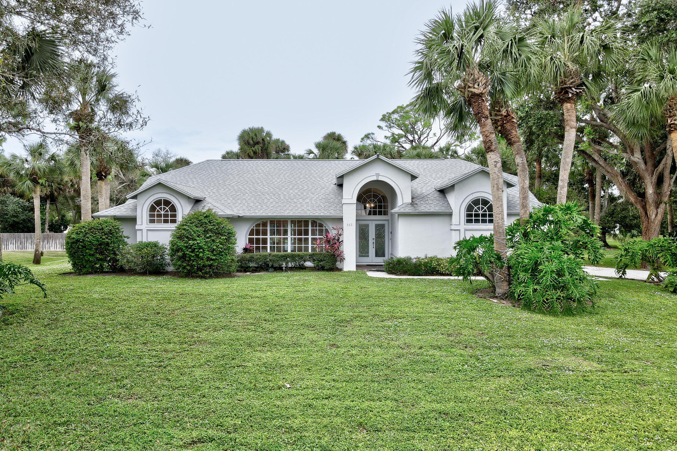 a view of a white house in front of a big yard with plants and large trees