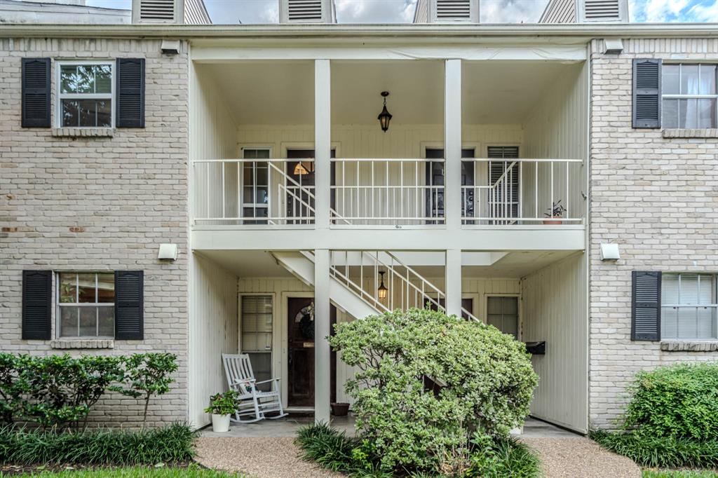 This is a two-story brick apartment building featuring a central staircase leading to upper-level units, each with a balcony. The ground floor units have individual entrances and there are green shrubs in front of the building.