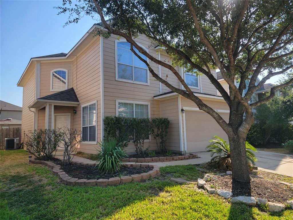 This is a two-story suburban home with beige siding, featuring a two-car garage, large windows, and a well-maintained lawn and landscaping.