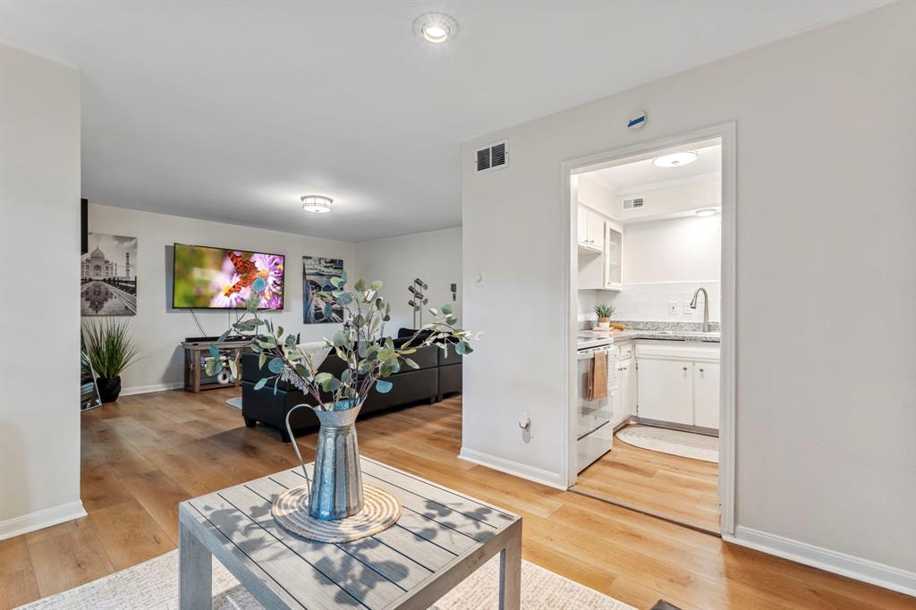 Modern living room with hardwood floors, a flat-screen TV, a couch, artistic wall decor, and an adjoining kitchen visible through a doorway.