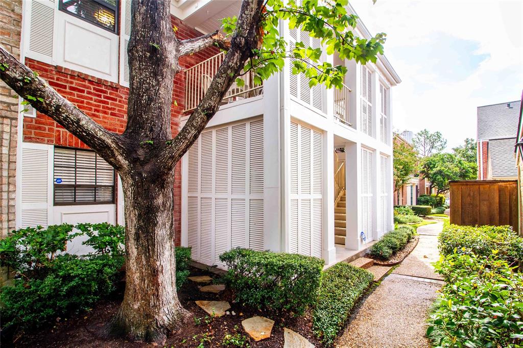 This is a charming multi-story brick townhouse with white accents, featuring a welcoming entrance flanked by plantation shutters and a mature tree providing shade to the facade. The property includes manicured shrubbery lining a stone pathway, creating a cozy and inviting atmosphere.