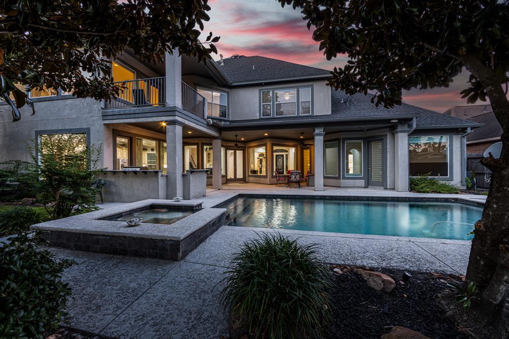 This is a two-story modern home at dusk featuring an inviting outdoor pool, a hot tub, and a well-appointed patio area, complemented by mature landscaping.