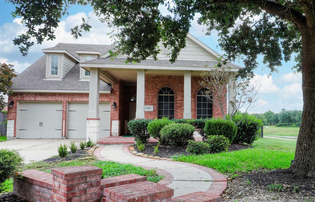 This is a single-story brick home featuring a covered entryway, arched windows, a two-car garage, and a manicured front lawn with a pathway leading to the front door. It's situated in a pleasant neighborhood with green surroundings.