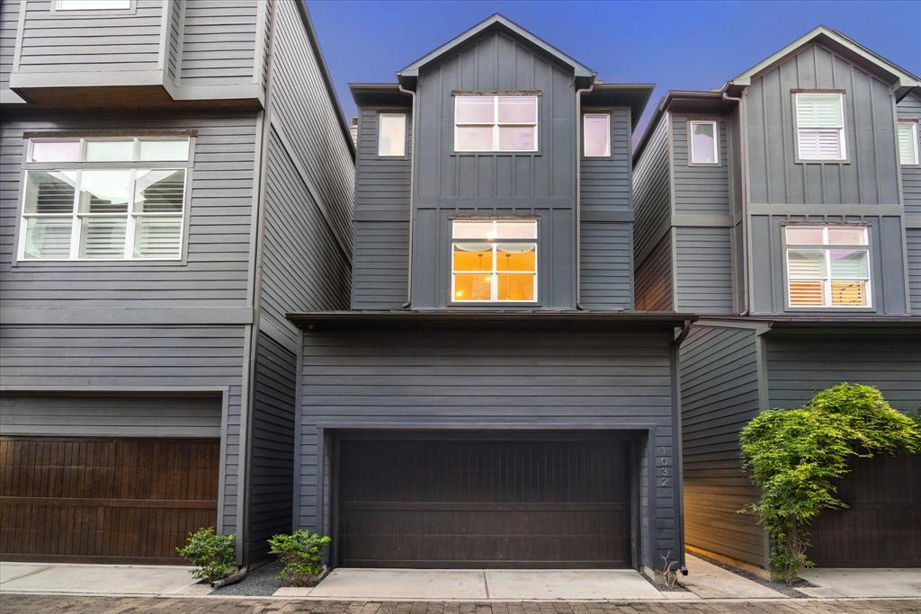 This home features modern, multi-story townhouse with dark gray exterior siding and lots of windows to provide natural light. The residence features a two car garage with wooden panels and greenery located near the entrance.