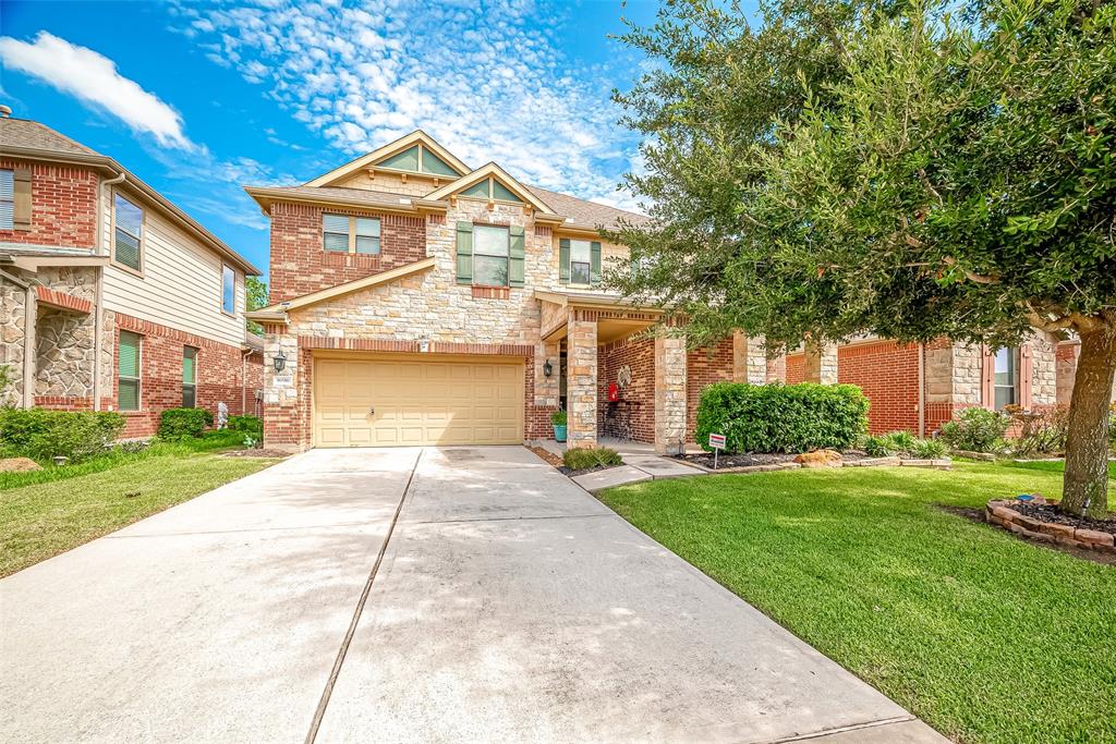 This is a two-story brick and stone facade home featuring a nicely manicured lawn, mature trees, and an attached two-car garage. The property is located in a suburban neighborhood with similar style houses.