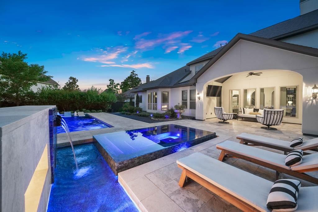 This elegant outdoor living space features a sleek infinity pool with a spa, surrounded by a well-appointed patio and lounge area. The home's exterior displays a modern design with arching doorways and a covered seating area that promises comfort and luxury.