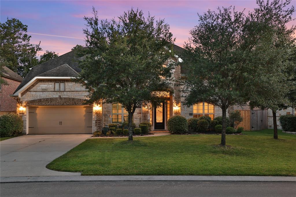 This is a two-story brick home with an attached two-car garage, well-manicured lawn, and mature trees, captured at dusk with exterior lighting on.