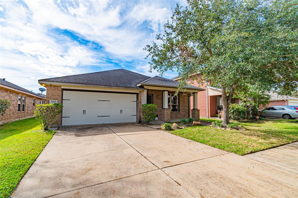 This is a single-story brick home featuring a two-car garage, a well-maintained lawn, and a mature tree in the front yard, situated in a suburban neighborhood.