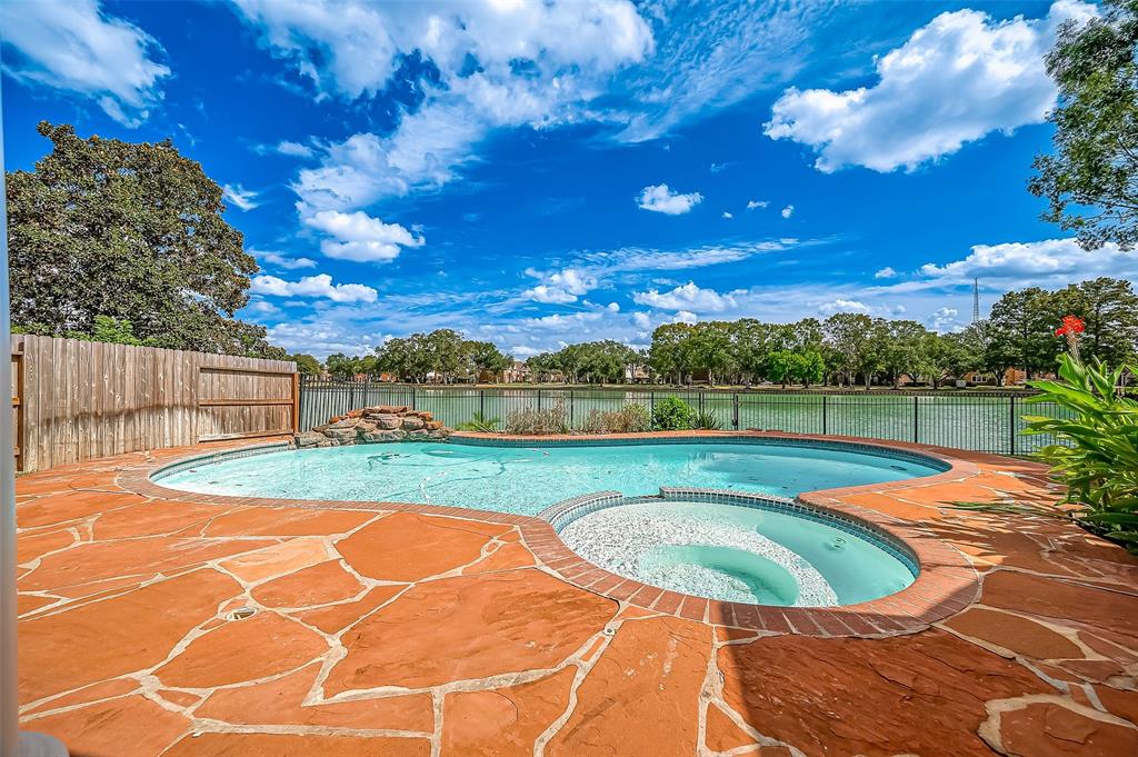 This photo showcases a spacious outdoor area with an inviting swimming pool, complemented by a beautiful lake front view and a clear blue sky. The poolside features a patterned stone deck, and the property is bordered by a wooden fence for privacy.