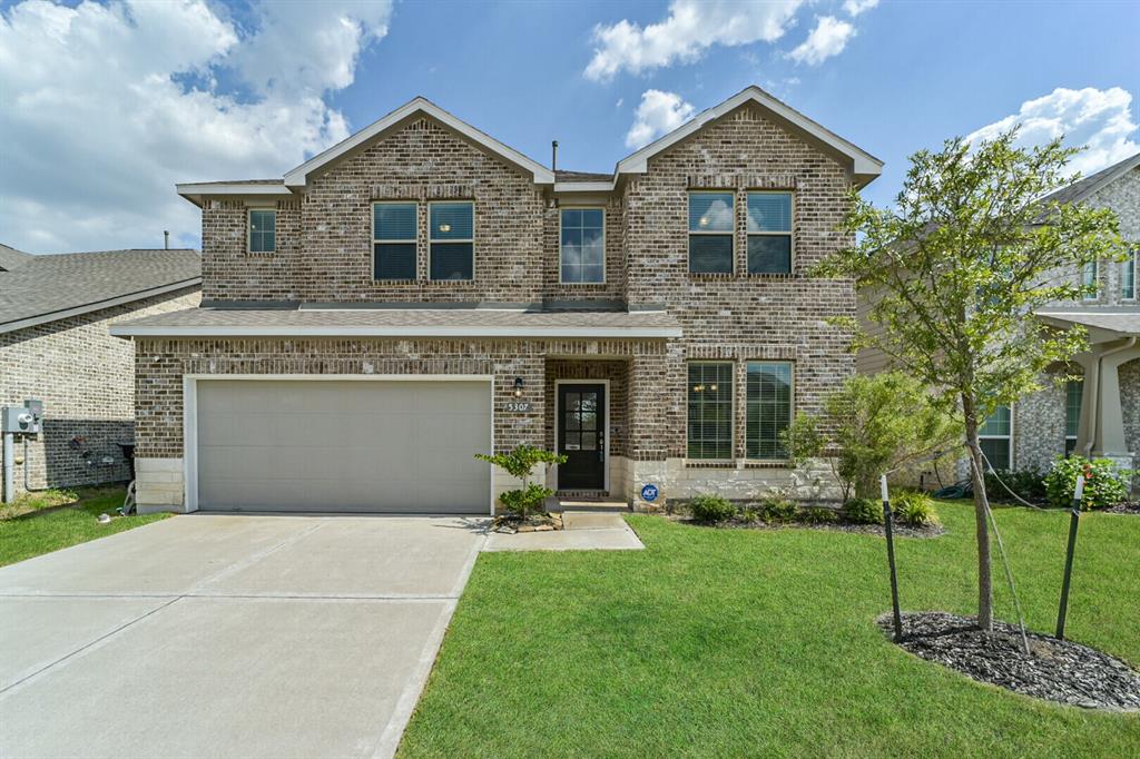 This is a two-story brick home featuring a two-car garage, a well-maintained lawn, and a young tree in the front yard. The brick and stone combination give this home a contemporary luxury feel.