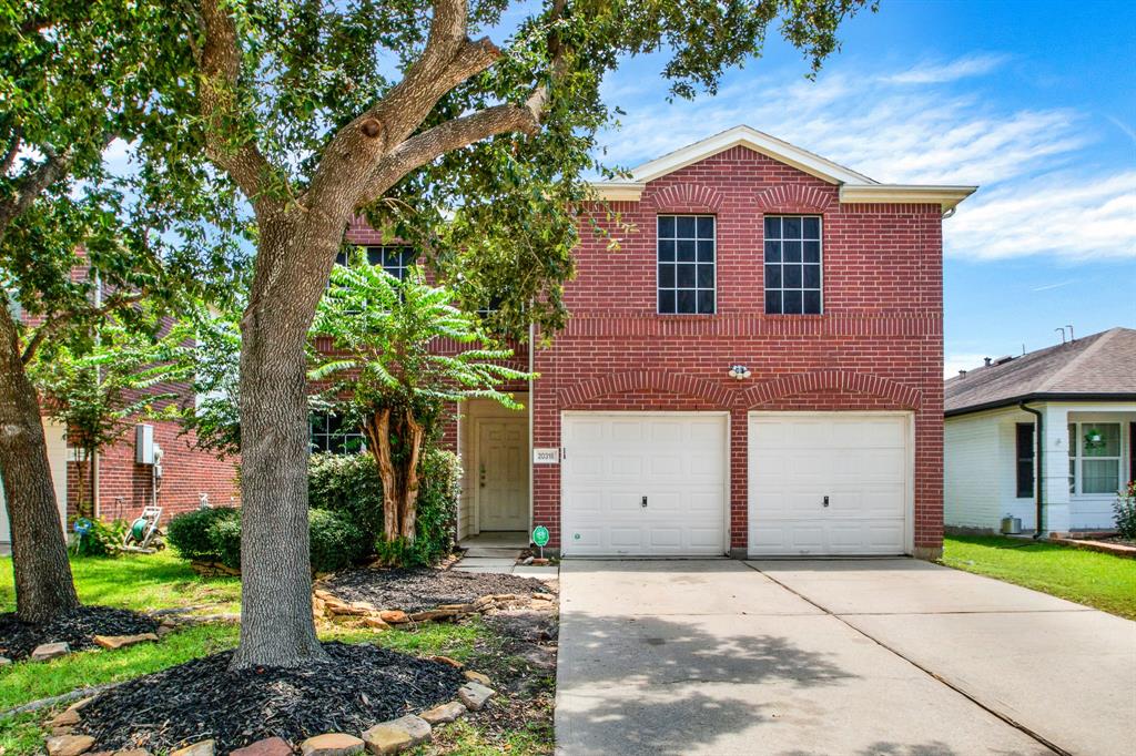 Two-story red brick house with a white door, double garage doors, a well-kept lawn, and a large shade tree in the front yard.