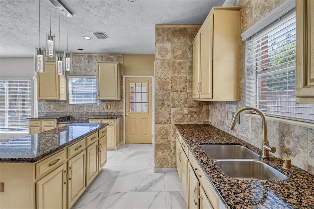 This is a spacious kitchen featuring light-colored cabinetry, granite countertops, modern hanging lights, and tiled backsplash and walls. The room is well-lit with natural light from large windows and has a tile floor.