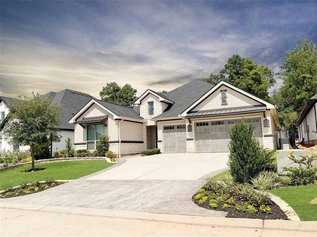 This is a modern single-story home featuring a clean, neutral color palette, a three-car garage, and large windows for natural light. The landscaping includes young trees and a well-maintained lawn, nestled within a tranquil, wooded setting.