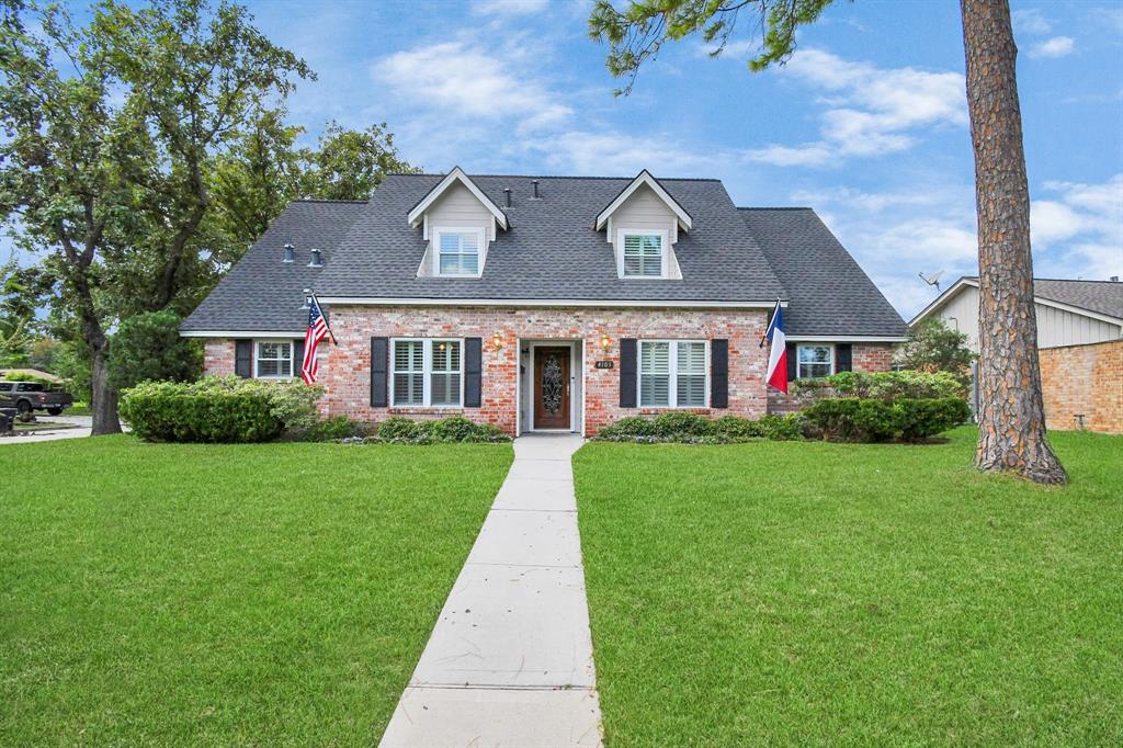 This is a charming two-story brick home featuring dormer windows, a well-manicured lawn with mature trees, and a welcoming walkway leading to a red front door flanked by American and Texas flags.