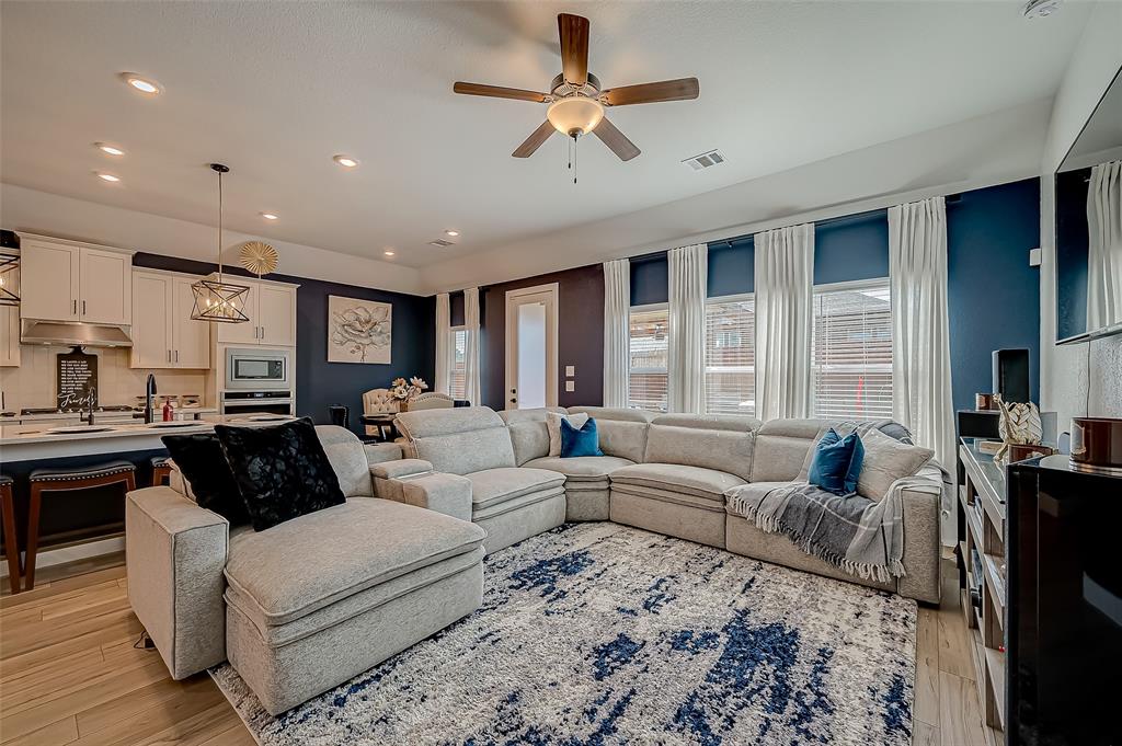 This photo shows a modern, open-concept living space with a comfortable sectional sofa, a well-equipped kitchen with white cabinetry, and a cozy dining area. The room is finished with stylish accents and a neutral color palette with blue tones, providing a perfect blend of function and elegance for contemporary living.