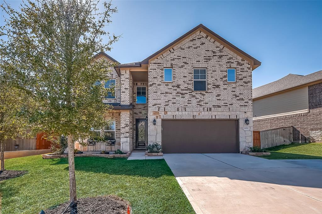 This is a two-story home with a brick exterior, featuring a two-car garage, and a neatly landscaped front yard with a young tree. The house has blue window shutters adding a pop of color.