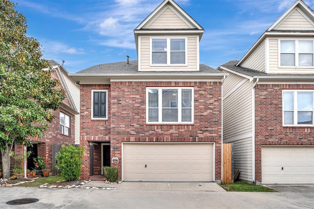 This is a modern, three-story townhouse with a brick exterior and a two-car garage. It's nestled in a row of similar homes, featuring a clean, well-maintained facade and a small front garden area.