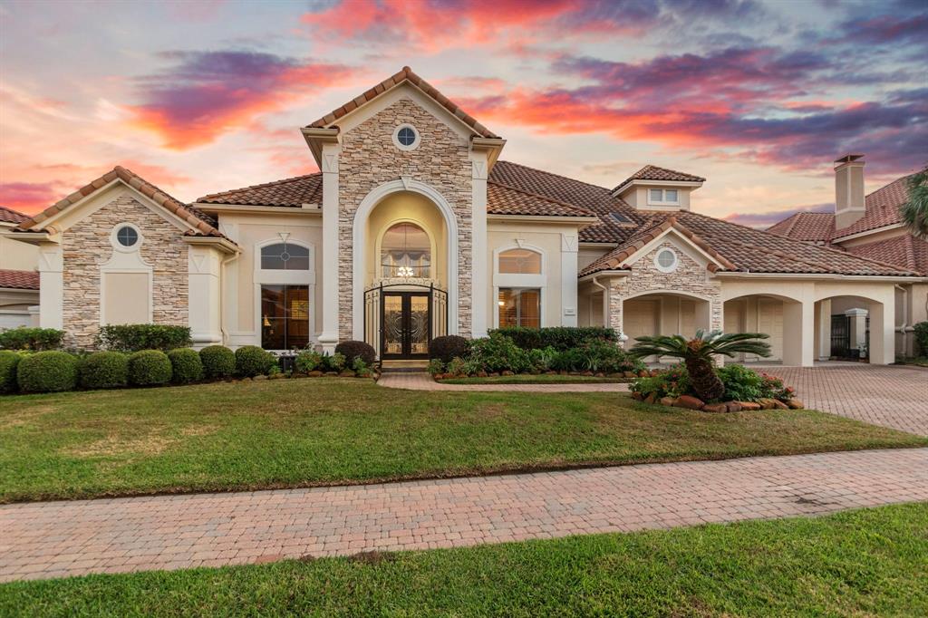 This is a spacious two-story home featuring a stone and stucco facade with a grand arched entrance, manicured landscaping, and a picturesque sunset backdrop.