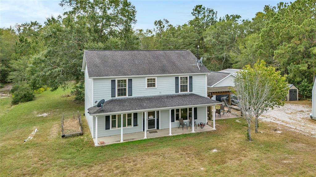 This is a two-story home with a gray exterior and white trim, featuring a covered front porch and an attached carport. The property is surrounded by greenery and trees, offering a sizable yard and a private setting.