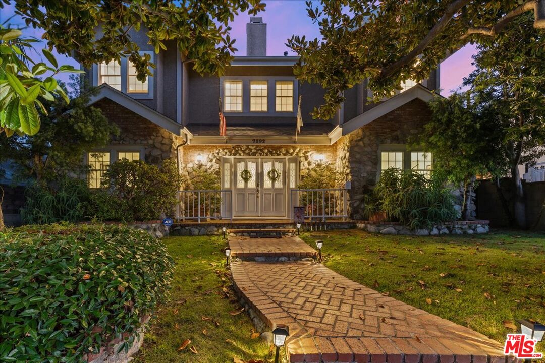 a view of a brick house with many windows plants and large trees