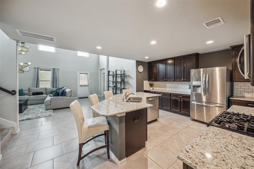 Modern open-concept kitchen and living area. The kitchen features granite countertops, a large island with seating, stainless steel appliances, and dark wood cabinets. The adjacent living space is bright with high ceilings and large windows, creating a welcoming atmosphere.