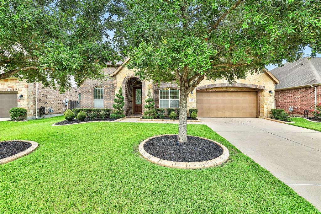 This is a single-story brick home with an arched entryway, manicured lawn, mature trees, and a two-car garage, presenting a well-maintained curb appeal in a suburban neighborhood.