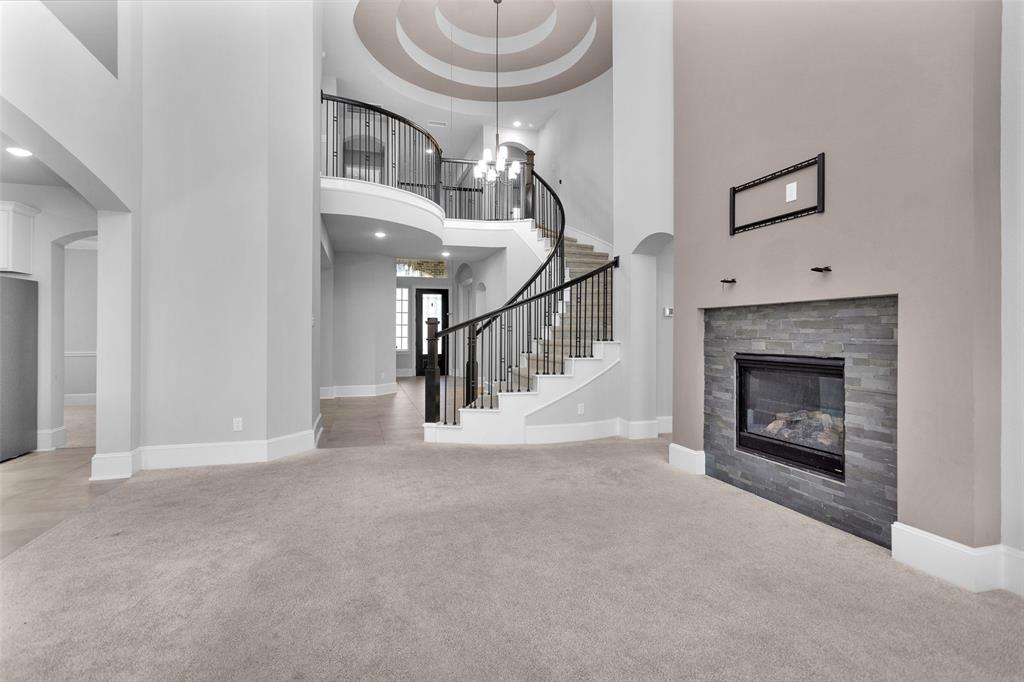 This image shows a spacious living area with high ceilings, featuring a modern fireplace with a stone surround, plush carpeting, and an elegant curved staircase with wrought-iron balusters leading to a second-floor balcony. The neutral color palette offers a versatile backdrop for personalization.