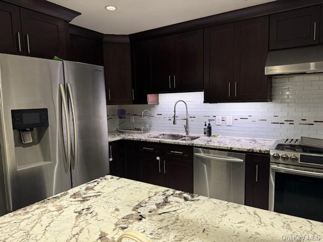 Kitchen featuring sink, decorative backsplash, light stone countertops, dark brown cabinets, and stainless steel appliances