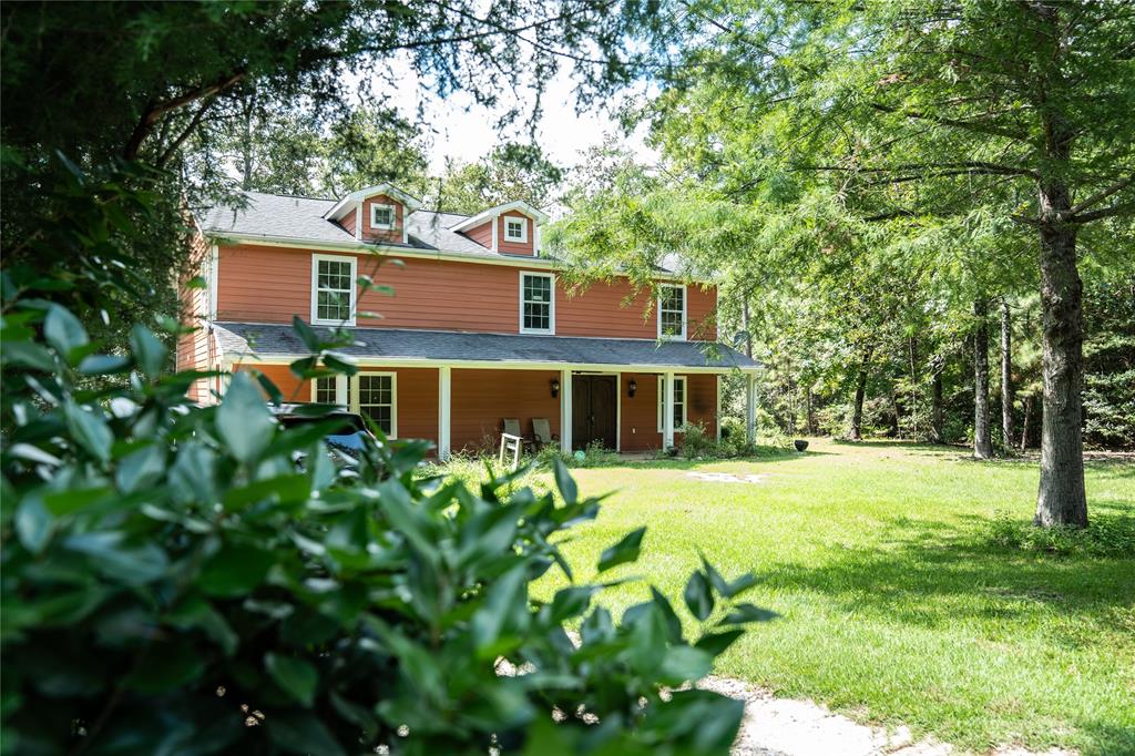 This photo shows a two-story home with a reddish exterior and white trim. It features dormer windows, a covered porch, and is nestled among mature trees in a lush, green yard, giving it a secluded and natural setting.