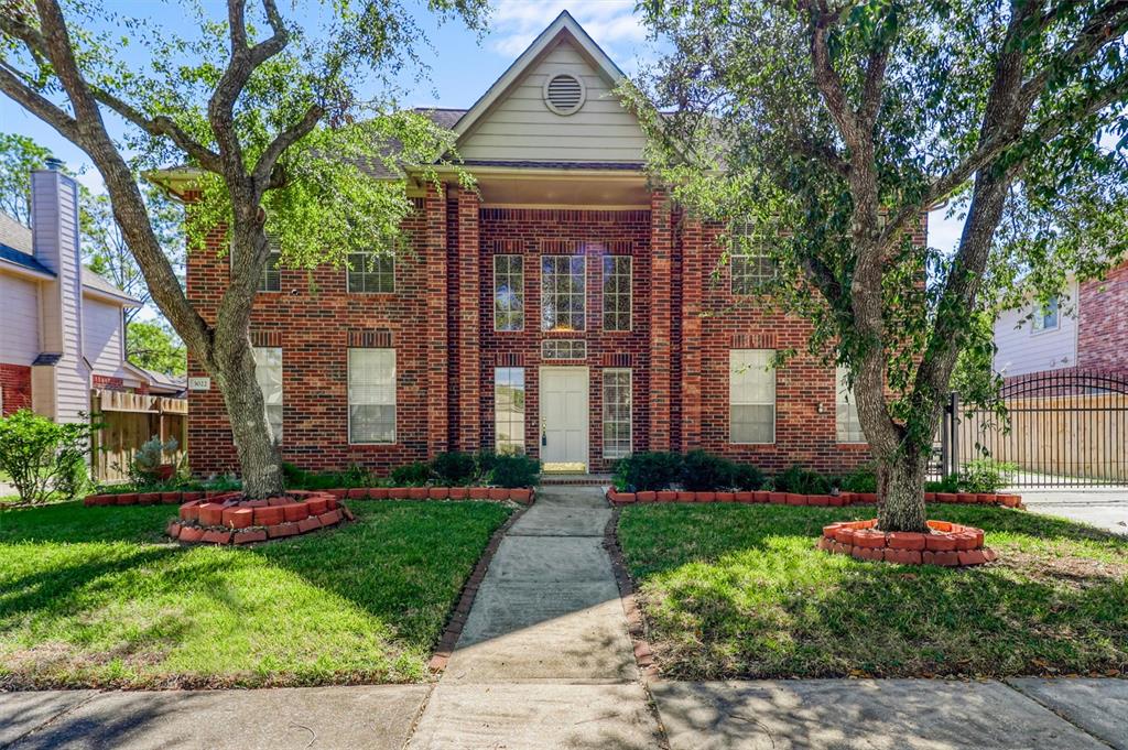 This is a two-story brick house with a symmetrical facade, featuring a central doorway with a transom window, flanked by two large windows on each side. It is nestled between mature trees, has a manicured lawn, and a concrete pathway leading to the entrance. The property is fenced on one side and appears to be in a suburban setting.