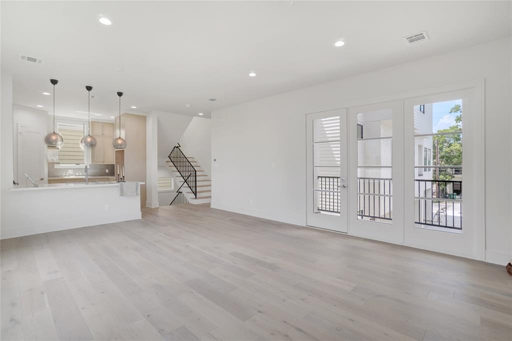 Open-concept living space with a combined kitchen area, featuring light hardwood floors, white cabinetry, and sleek pendant lighting. The room is well-lit with natural light from French doors leading to a balcony and boasts a contemporary staircase with black accents.