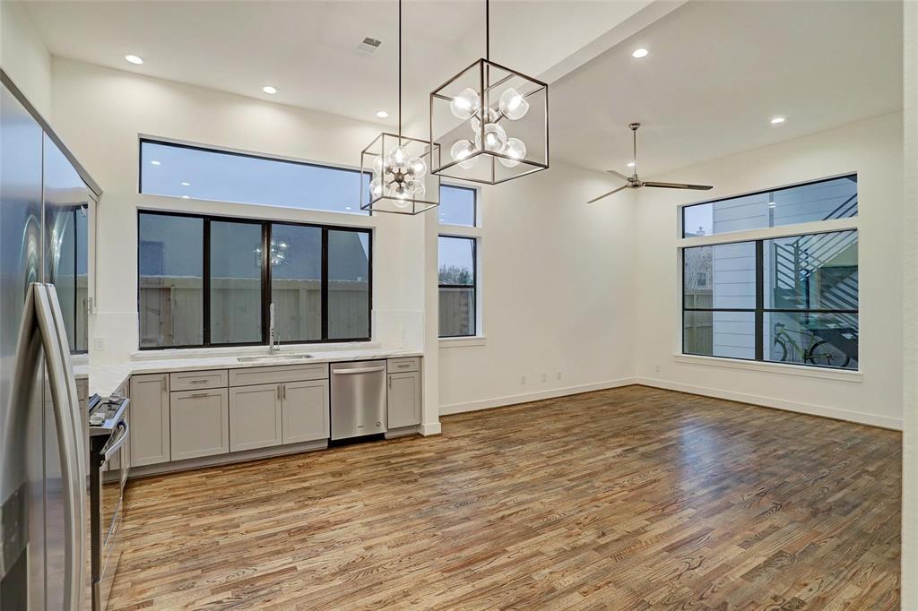 This is a modern, open-concept living space with high ceilings, large windows for natural light, a contemporary lighting fixture, ceiling fan, and a sleek kitchenette with stainless steel appliances. The room features hardwood flooring and a neutral color palette.