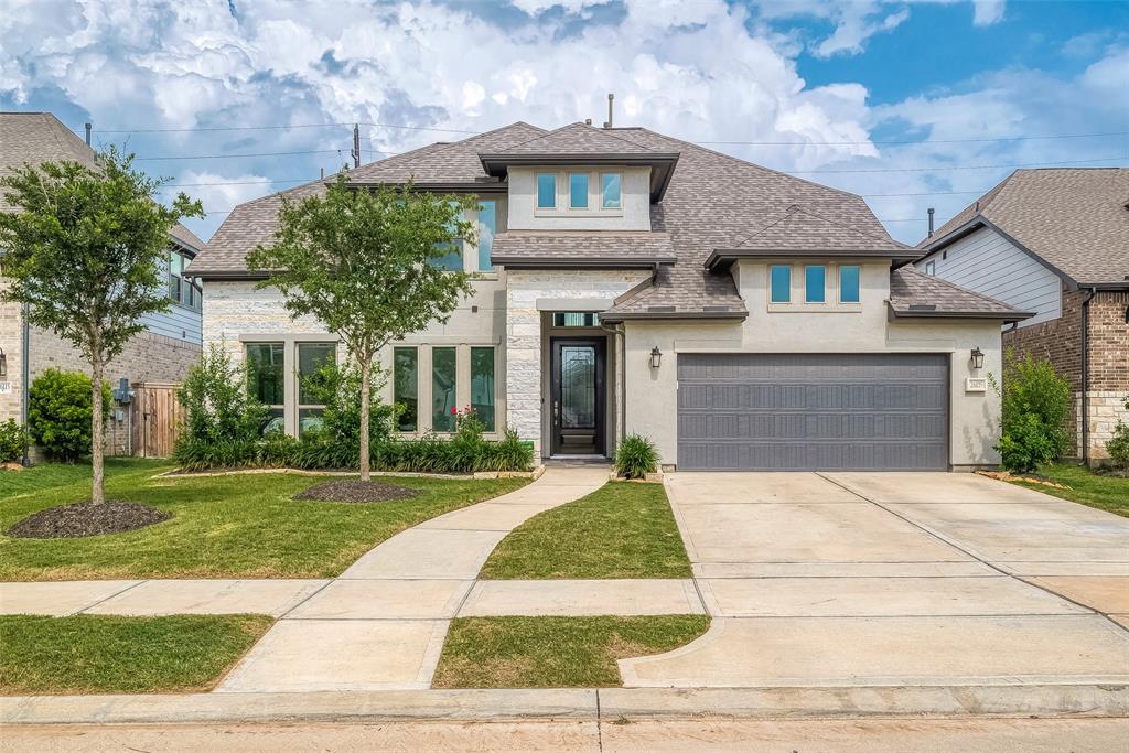 This is a modern two-story home featuring a mix of stone and stucco exterior, a two-car garage, and a landscaped front yard with young trees. The house has large windows and a covered front entryway.