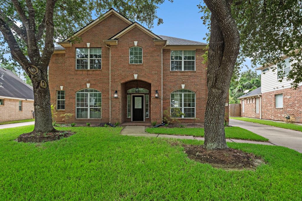 Welcome home! Two-story brick home featuring large windows, an arched entryway, and is shaded by mature trees.