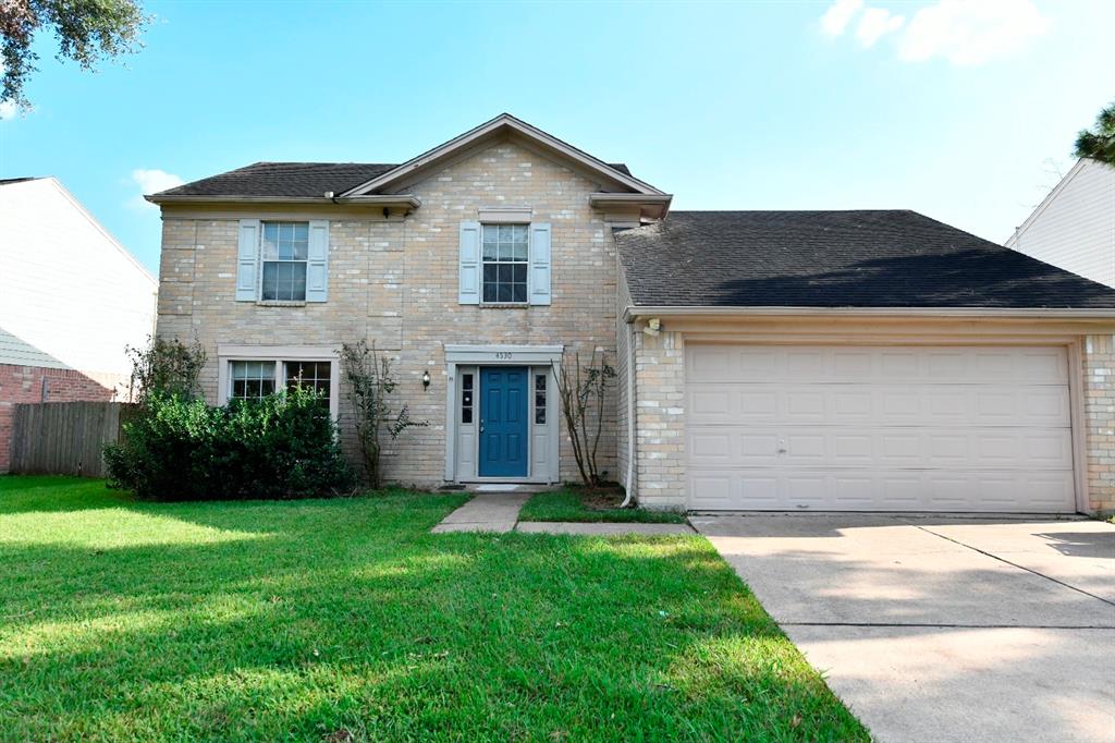 This is a two-story, traditional-style home featuring a tan brick exterior, an attached two-car garage, and a blue front door. The house has a well-maintained lawn and is situated in a suburban neighborhood.