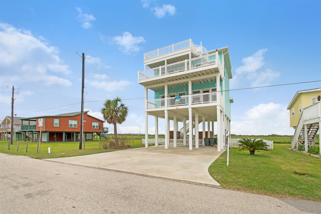 This is a multi-story, elevated beach house with a pale blue exterior, extensive balconies on each level, and ample under-house parking. 