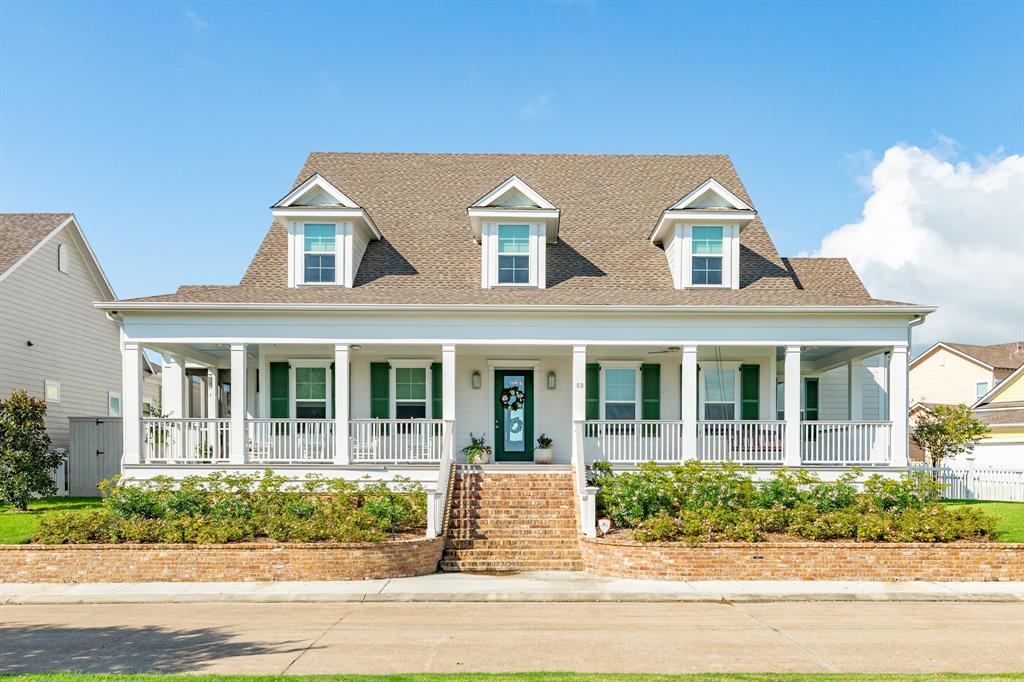 Charming two-story home with a welcoming front porch, dormer windows, and a traditional peaked roofline. It features a well-maintained lawn and an attractive brick-lined flower bed in the front.