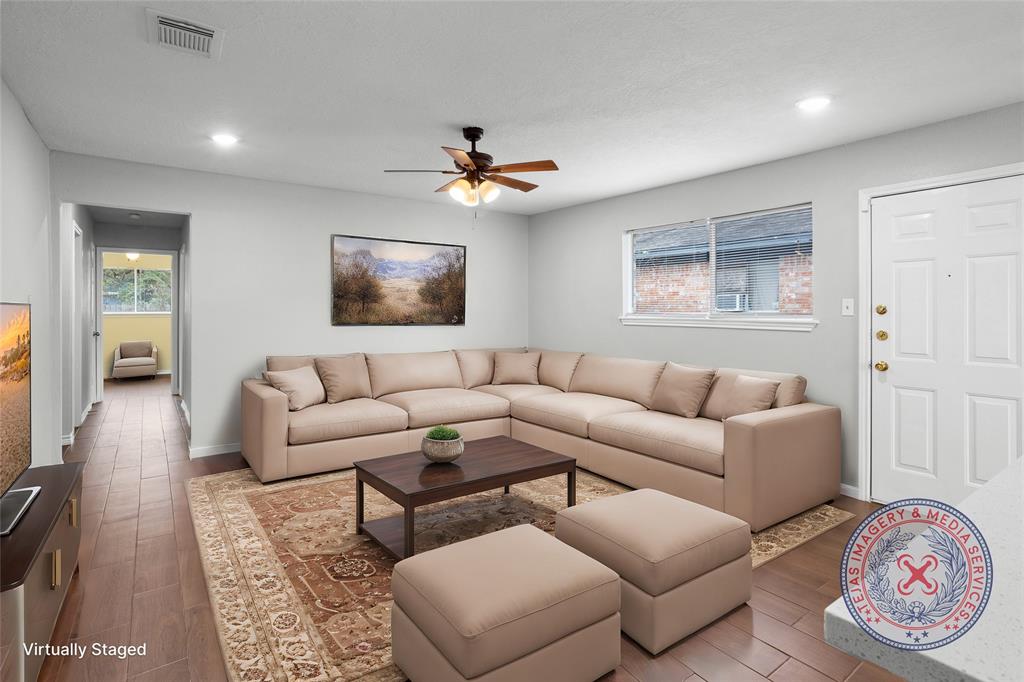 This photo shows a bright, open living room with wood-look tile flooring, a ceiling fan, and a window providing natural light. The neutral walls and recessed lighting create a modern, inviting space.