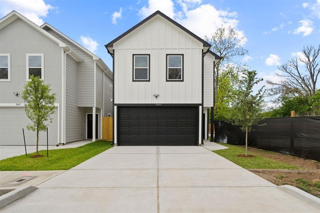 A modern two-story home with a clean white and black exterior. It features a spacious driveway leading to a black garage door, with a small lawn and young trees on either side. The house is part of a neat neighborhood setting.