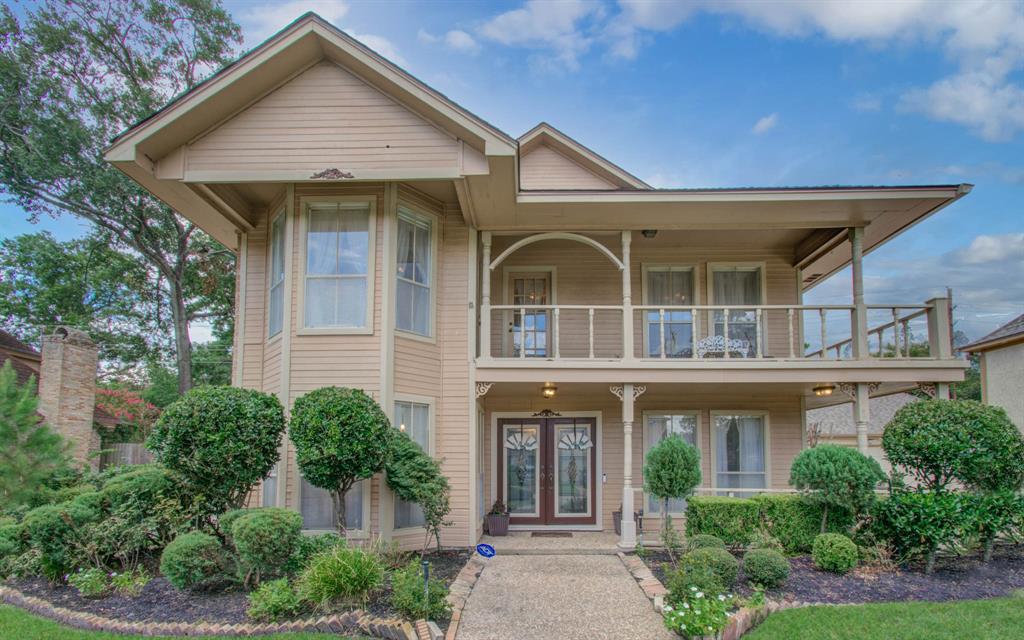 This is a two-story home with a beige exterior, featuring a prominent gable, large windows, and a welcoming front porch with a balcony above. The property is well-landscaped with manicured shrubs and a tidy lawn.