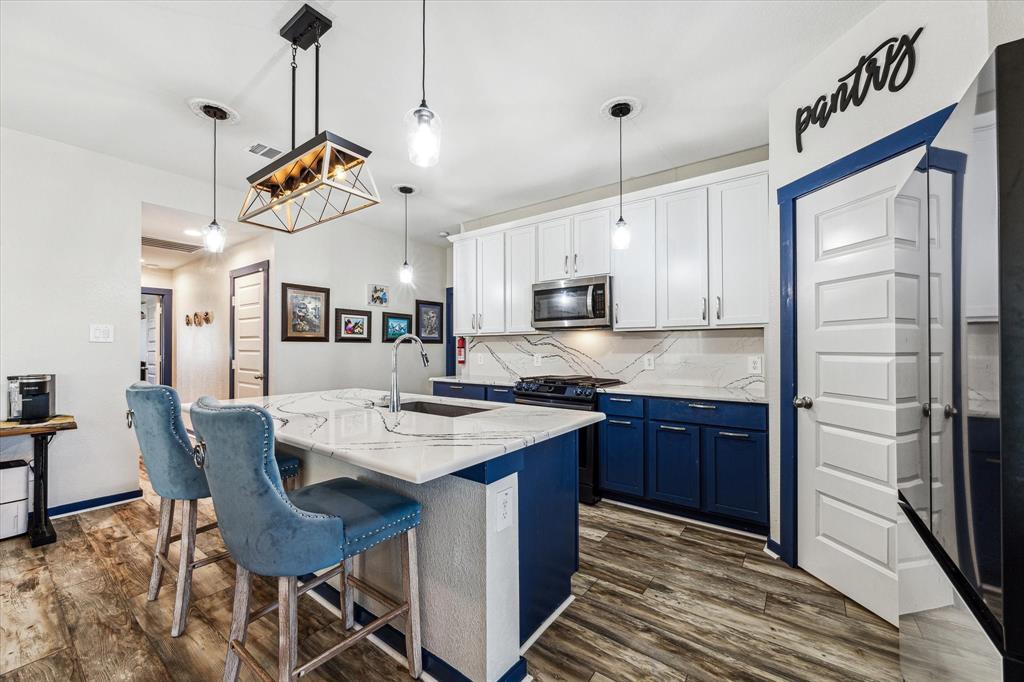 This is a modern, open-concept kitchen featuring white upper cabinetry and contrasting blue lower cabinets, with stylish pendant lighting above an island with bar seating. The kitchen also includes updated appliances and has a dedicated pantry space marked by a playful sign.