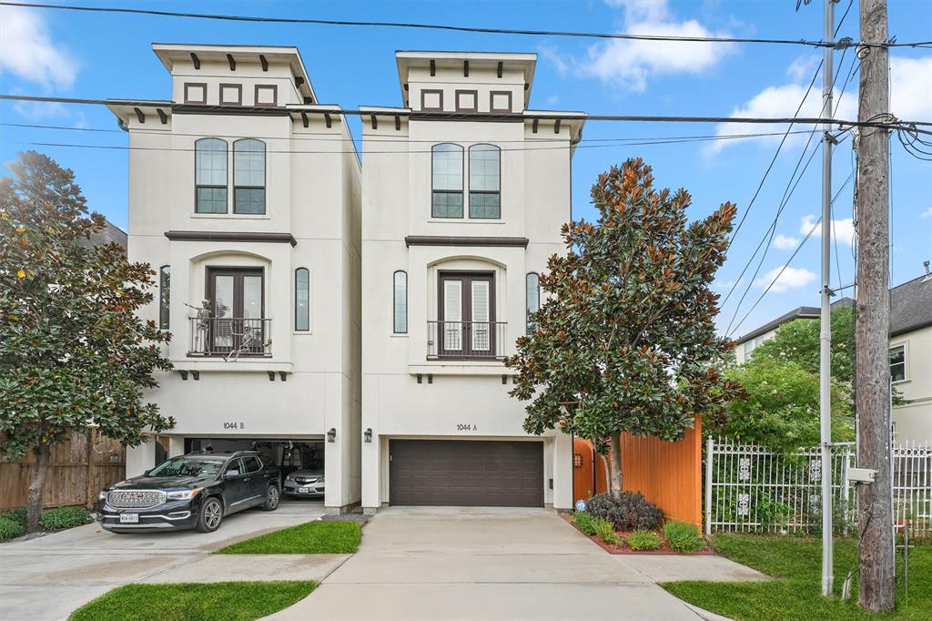 This is a modern, multi-story duplex with a light facade, featuring prominent architectural details and Juliet balconies. an attached garage and the property includes a fence for privacy. Tall trees and greenery enhance the urban setting.