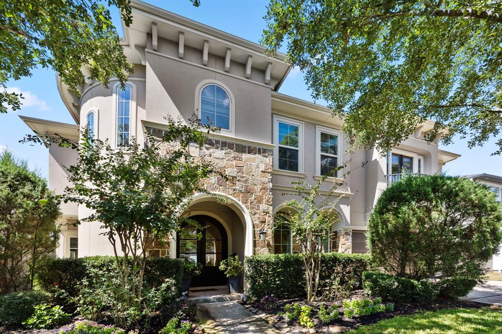 This is an elegant two-story home featuring a stone and stucco exterior, arched entryway, and large windows nestled amongst mature trees and well-maintained landscaping.